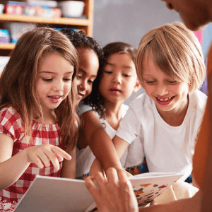 Picture of four children smiling and pointing at a book being held up in class.